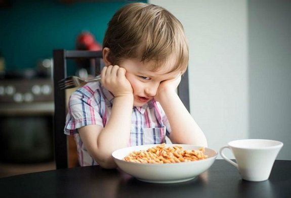 Маленький ребенок сидит за столом подпирая голову руками, перед ним тарелка с едой